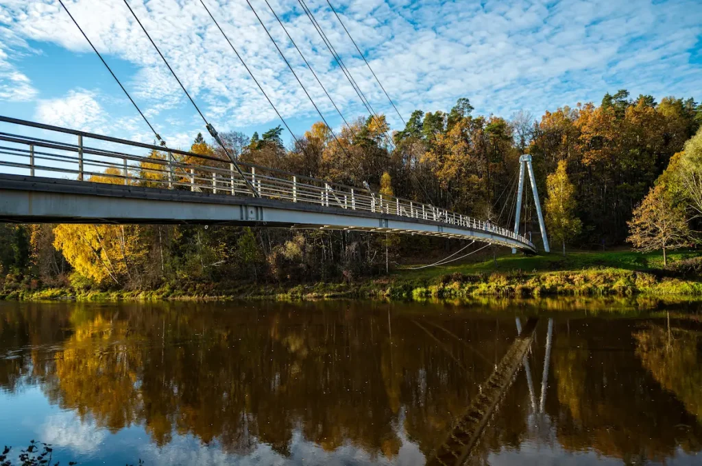 Modern suspension bridge over calm river with autumn trees, blue sky reflections, and peaceful natural scenery