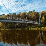 Modern suspension bridge over calm river with autumn trees, blue sky reflections, and peaceful natural scenery